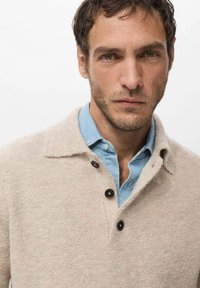 Beżowy sweter z wełny z kołnierzem, ozdobiony trzema czarnymi guzikami oraz widoczną niebieską koszulą dżinsową pod spodem. Miękka tekstura, neutralny kolor.