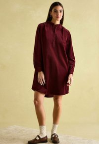 Burgunderfarbendes Cordkleid mit langen Ärmeln, Knopfleiste und seitlichen Schlitzen, kombiniert mit braunen Slippern und gepunkteten Socken. Strukturierter Stoff.