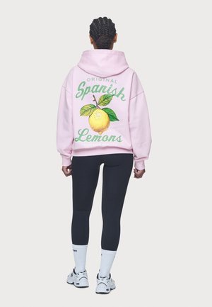 Persoon met ingevlochten haar, een roze hoodie met een citroengrafiek en de tekst "Original Spanish Lemons" op de achterkant, zwarte leggings, witte sokken en sneakers.