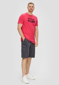 Rode katoenen T-shirt met "ACAPULCO" grafiek, gecombineerd met donkergrijze cargoshorts met zijzakken. Model draagt witte sneakers.