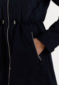Marine bleu gewatteerde jas met een getailleerde taille, ritssluitingen en een gladde stoftextuur. Zilverkleurige ritsen contrasteren met de donkere kleur.