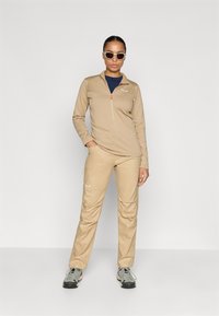 Μπεζ φούτερ με φερμουάρ και μακριά μανίκια, συνοδευόμενο από παντελόνι που ταιριάζει, κατασκευασμένο από υφασμάτινη επιφάνεια. Φορεμένο με γυαλιά ηλίου και ανθεκτικά υποδήματα. Μινιμαλιστικές σχεδιαστικές λεπτομέρειες.