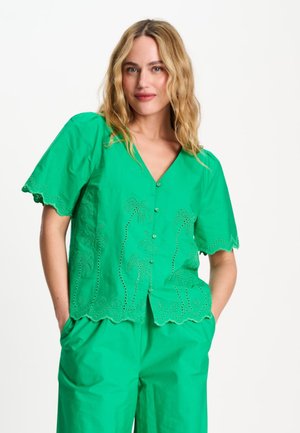 Vrouw met golvend blond haar die een groene blouse met knopen en palmboomuitstansingen draagt en bijpassende groene broek, handen in de zakken, neutrale achtergrond.