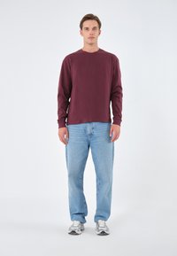 Weinrotes langärmliges Hemd, kombiniert mit hellblauen Jeans. Das Hemd hat einen runden Halsausschnitt, und der Mann trägt silberne Sneakers.