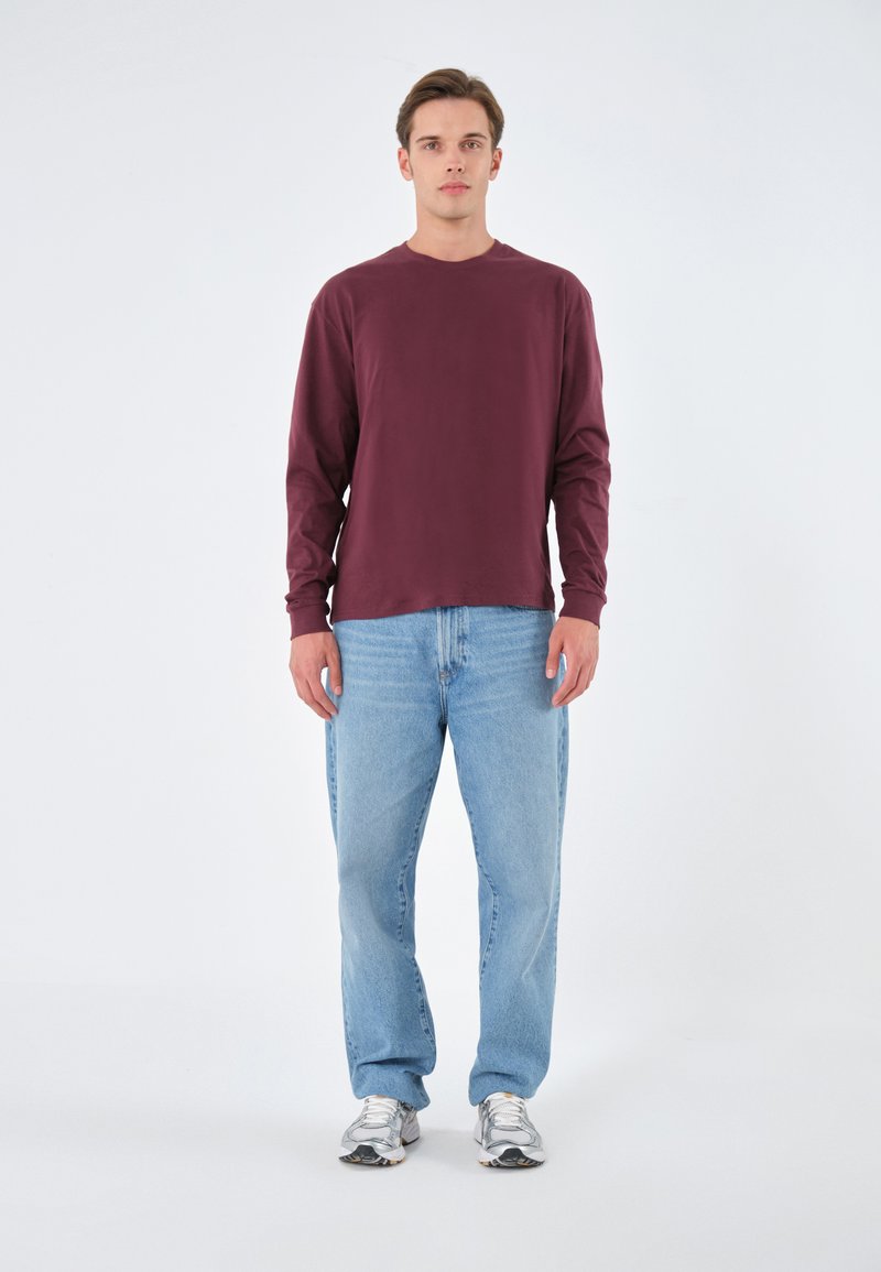 Weinrotes langärmliges Hemd, kombiniert mit hellblauen Jeans. Das Hemd hat einen runden Halsausschnitt, und der Mann trägt silberne Sneakers.
