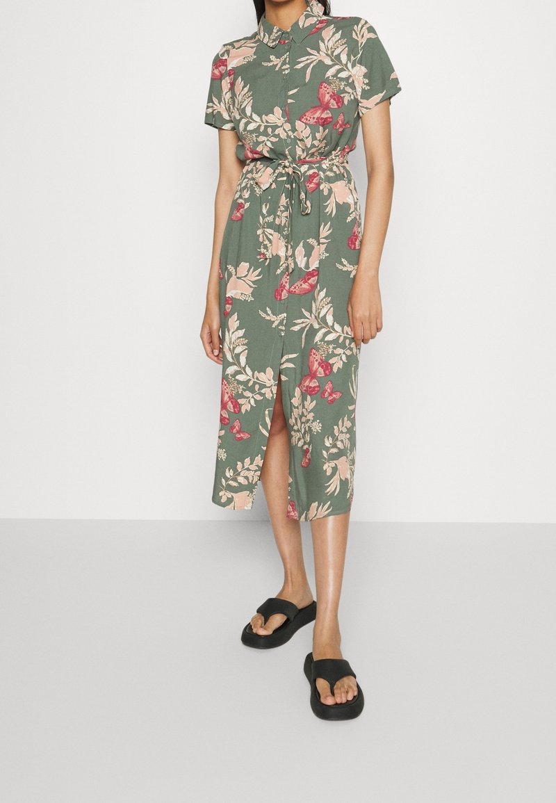 Femme portant une robe chemise midi verte avec des imprimés floraux roses et papillons, nouée à la taille, associée à des sandales noires ouvertes à l'avant.