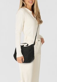 Borsa nera trapuntata con trama a righe diagonali, tracolla regolabile e forma curva. Indossata a spalla, abbinata a una camicia bianca con colletto.