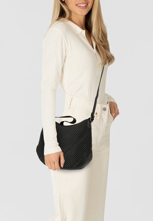 Zwarte gewatteerde tas met diagonale ribbeltextuur, verstelbare schouderriem en een gebogen vorm. Draagt op de schouder, gecombineerd met een wit overhemd met kraag.