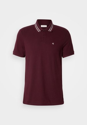 Meeste tumepunane lühikeste varrukatega polo särk, millel on kahe nupuga kaelus, triibuline kaelaäär ja väike valge "cK" logo rinnal.