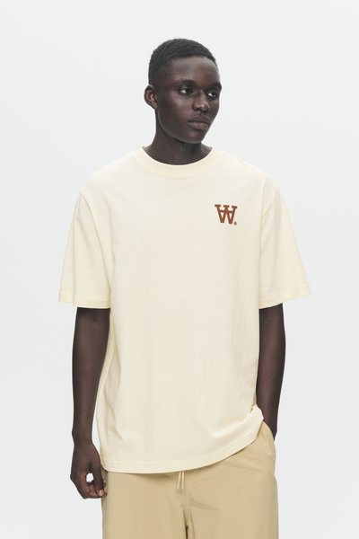 Giovane indossante una t-shirt oversize crema con un piccolo logo marrone a "W" sul petto e pantaloni corti beige, in piedi contro uno sfondo bianco.