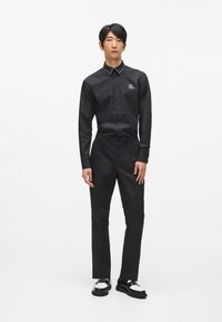 Melna krekla ar pogām apkakli, kombinēta ar pielāgotām melnām biksēm un melnbaltiem apaviem. Gluda auduma, minimālisma dizains.