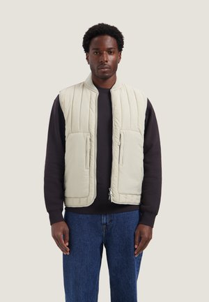 Uomo che indossa un gilet trapuntato beige chiaro sopra una maglia nera a maniche lunghe e jeans blu, in piedi davanti a uno sfondo chiaro e uniforme.