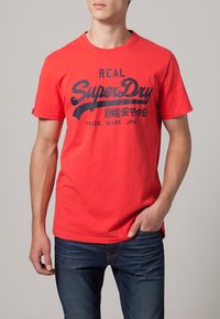Camiseta de algodón roja con mangas cortas, que presenta un logo y texto impresos en negro en el frente. Combinada con jeans azules.