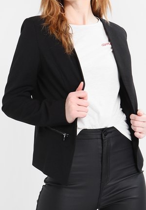 Kvinde iført en sort blazer over en hvid t-shirt og sorte bukser, der holder blazerens kanter med begge hænder.