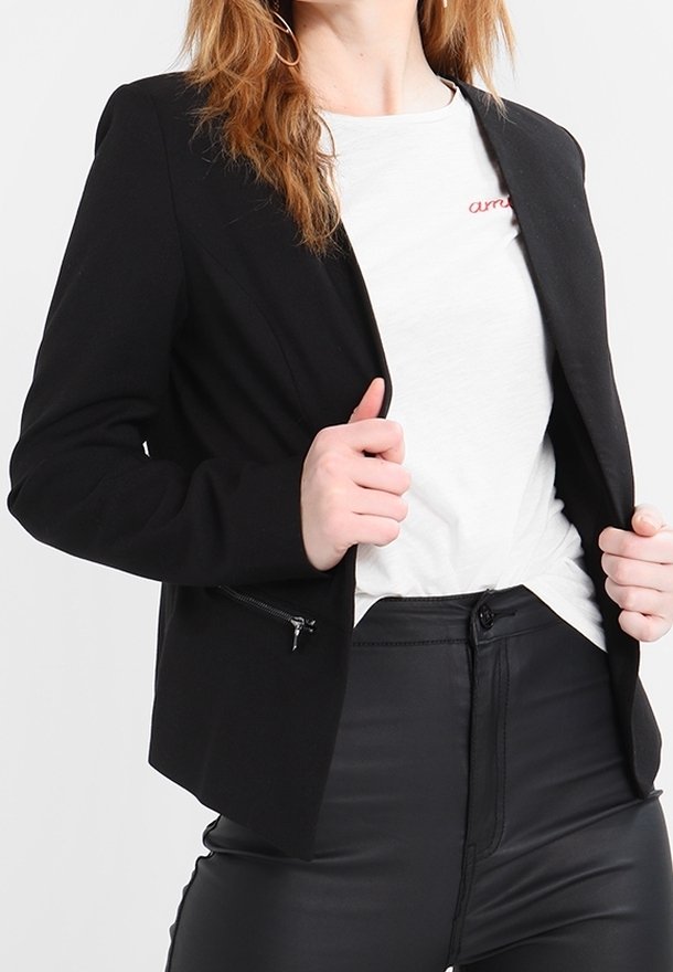 Femme portant un blazer noir par-dessus un t-shirt blanc et un pantalon noir, tenant les bords du blazer avec les deux mains.