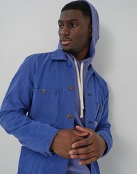 Blue corduroy jacket with a collared design, featuring two front pockets and hardware buttons, layered over a light gray hooded sweatshirt.