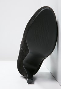 Zapato de tacón alto de ante negro visto desde atrás, con punta afilada y suela de goma texturizada con ligeras estrías de tracción.