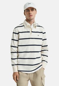Witte polo met lange mouwen en marineblauwe strepen, voorzien van een knoopsluiting bij de kraag en een geborduurd logo op de borst. Draagt bij beige broek.