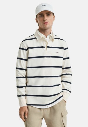 White long-sleeve polo shirt with navy stripes, featuring a buttoned collar and embroidered logo on the chest. Worn with beige pants.