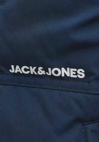 Tumši zila auduma daļa ar baltu "JACK & JONES" logotipu, kas izvietots virs horizontālas šuves.