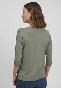 Pull en tricot vert à manches trois-quarts. Présente des poignets roulés et des fentes sur les côtés. Texture lisse et coupe décontractée.