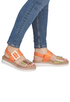 Feet wearing orange sandals with white buckles and multicolored front straps, paired with blue skinny jeans.