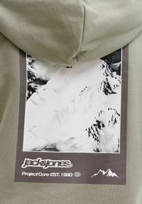 Obdĺžnikový grafický motív zasnežených horských vrcholov na šedej látke s logom "Jack & Jones" a textom "Project Core EST. 1990" pod ním.