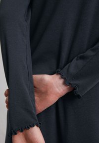 Ciemnoszara bluzka z długim rękawem i falbaniastymi mankietami. Gładki materiał, dopasowany krój, z ręką umieszczoną z tyłu. Prosta i czysta estetyka.