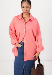 Chemise corail oversize à boutons, avec col et manches retroussées, associée à une jupe en denim et un sac tote marron. Texture de tissu lisse.