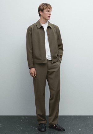 Young man standing, wearing olive green zip-front jacket and matching pants, white T-shirt, and black closed-toe sandals against pale wall.