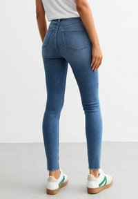 Jeans de denim azul ajustados con cintura alta y dobladillo deshilachado, combinados con zapatillas blancas con un acento verde. Dos bolsillos traseros visibles.