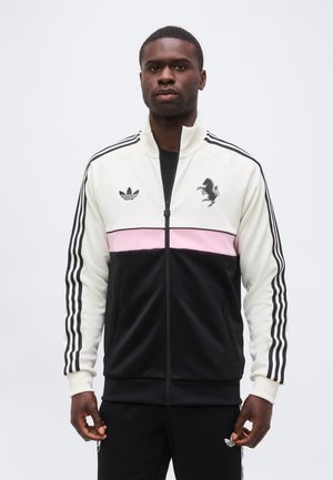 Uomo che indossa una giacca da allenamento Adidas bianca, rosa e nera con strisce nere sulle maniche e loghi sul petto, in piedi davanti a uno sfondo semplice.