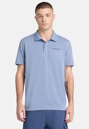 Junger Mann mit kurzen Haaren, der ein hellblaues Timberland-Poloshirt und dunkelblaue Cargo-Shorts trägt, vor einem schlichten Hintergrund stehend.