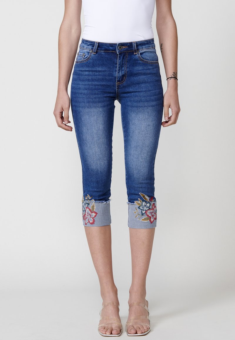 Jeans capri en denim bleu avec broderies florales aux poignets, taille mi-haute et coupe ajustée.