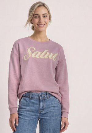 Smilende kvinde med blondt hår bærer en lyserød sweatshirt med "Salut" i gyldent skrift og blå jeans foran en ensfarvet lys baggrund.