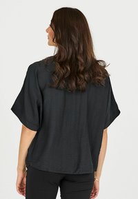 Schwarzes kurzärmeliges Blusenshirt mit strukturierter Oberfläche. Hat eine lockere Passform und seitliche Schlitze, von hinten gezeigt mit lockigem, welligem Haar.