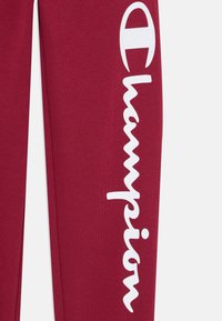 Pantalones de chándal color vino tinto hechos de una tela suave, presentan un gran logotipo blanco de "Champion" a lo largo de la pierna izquierda en una fuente estilizada.