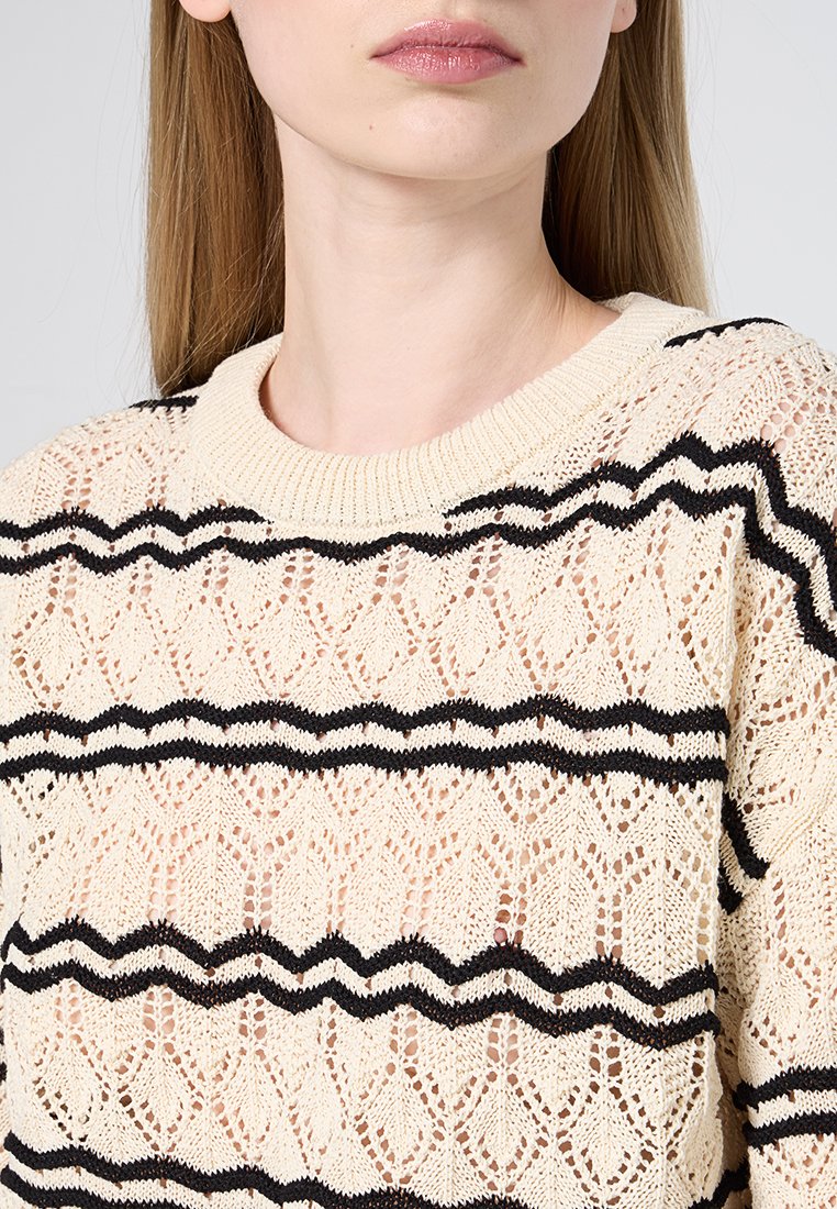 Crème gebreide trui met zwarte golvende strepen, met een kantachtige textuur en een geribde halslijn. Bovenlichaamsweergave benadrukt de ontwerpelementen.