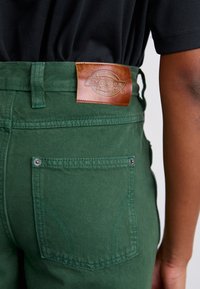 Green cotton pants featuring a leather brand patch, back pocket details, and contrast stitching. The pockets have metal snap closures.