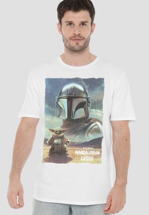 Mann trägt weißes T-Shirt mit dem Aufdruck des Mandalorian-Helms und Grogu, steht in der Wüste vor einem bewölkten Himmel.