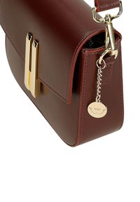 Bordeaux leren handtas met goudkleurige hardware, voorzien van een klepsluiting, een platte bodem en een decoratieve ketting met een ronde hanger.