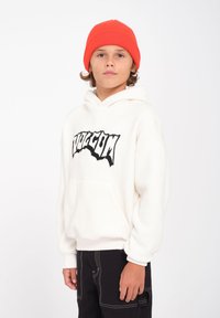 Witte fleece hoodie met zwart "Volcom" logo, voorzien van een kangoeroezak en ribgebreide manchetten, gecombineerd met zwarte broek en een oranje gebreide muts.