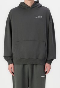 Sweat à capuche gris foncé en tissu doux, avec une poche kangourou et un logo blanc sur la poitrine. Coupe ample avec poignets et ourlet côtelés.