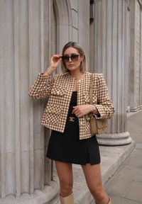 Geruite bruine en crèmekleurige jas met gouden knopen, gedragen over een zwarte jurk; geaccessoireerd met een donkere zonnebril en een beige handtas.