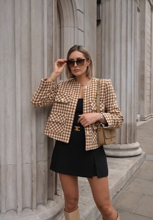 Veste à carreaux marron et crème avec des boutons dorés, portée sur une robe noire ; accessoirisée avec des lunettes de soleil foncées et un sac à main beige.
