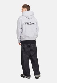 Grauer Kapuzenpullover mit einem großen schwarzen Logo auf dem Rücken, kombiniert mit dunklen Jeans und schwarzen Schuhen. Lockere Passform und schlichtes Design.
