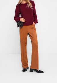 Pull en maille côtelée bordeaux associé à un pantalon large en tissu beige. Chaussures à talons noires à bout carré et petit sac à main noir.