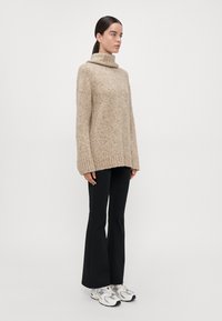 Beige gebreide trui met een hoge col, geribbelde zoom en manchetten, gecombineerd met zwarte flared broek en witte sportschoenen. Glad textuur.