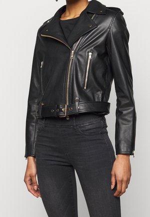 Persoon die een zwarte leren motorjas draagt met gouden ritsen en een taille met riem, gecombineerd met donkere high-waisted jeans.