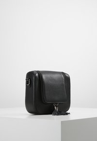 Borsa a tracolla in pelle nera con finitura texturizzata, frontale a patta, chiusura con zip e dettaglio in nappina. Presenta hardware in metallo e bordi arrotondati.